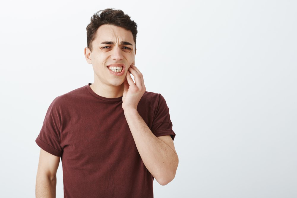 Portrait of displeased suffering young male model in red t-shirt, frowning and clenching fists, touching jaw while feeling toothache, having decay and dental problems, standing over gray background